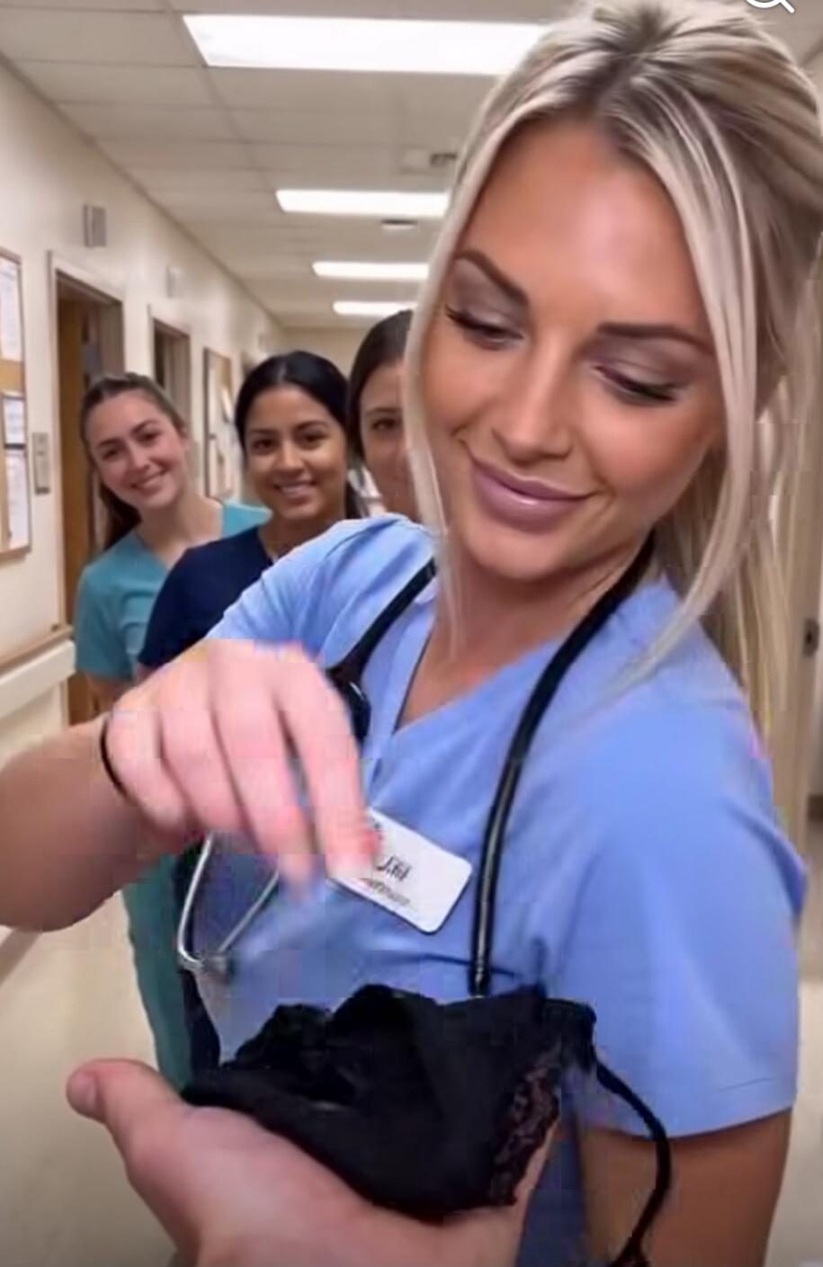 A woman in medical scrubs is holding up a pair of black lace underwear, smiling. Other women in scrubs are smiling behind her in a hospital hallway.