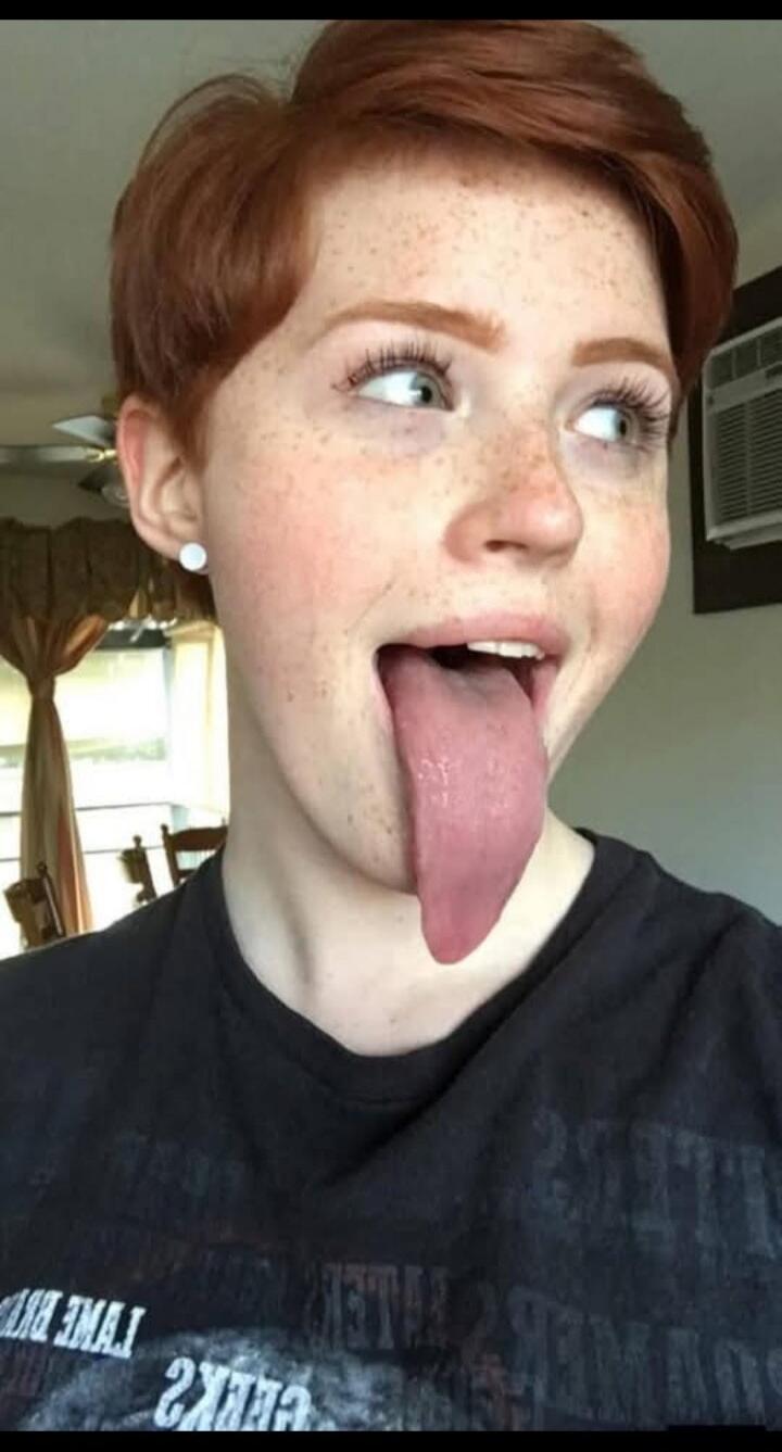 A person with short red hair and freckles is sticking out an unusually long tongue, looking upwards and to the side. The person is wearing a dark t-shirt with some text on it.