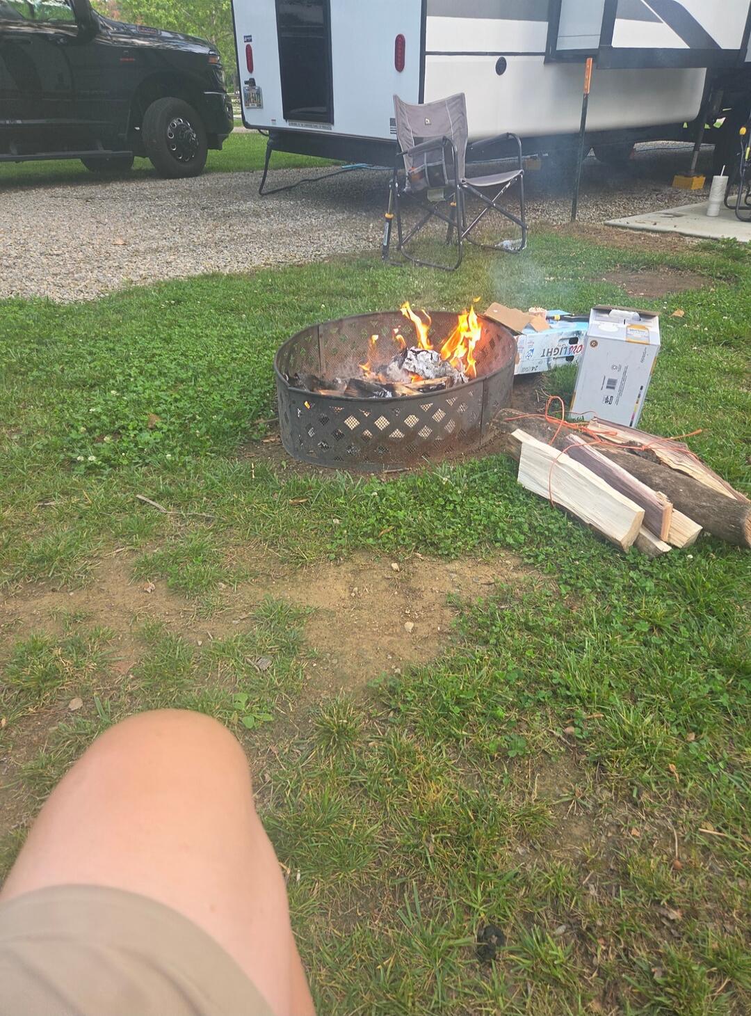 A campfire burning brightly in a metal fire pit on a grassy patch of ground. A stack of firewood is neatly placed beside the pit. In the background, a black pickup truck and a white recreational vehicle (RV) are parked, suggesting a campsite setting. A folding camping chair is visible near the RV. A box, partially visible, shows the branding 'COORS