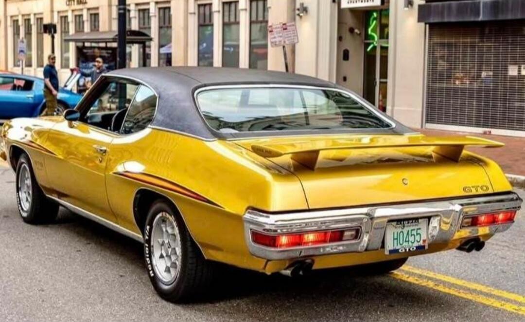 A classic yellow Pontiac GTO with a black roof is parked on a city street. Text visible includes 
