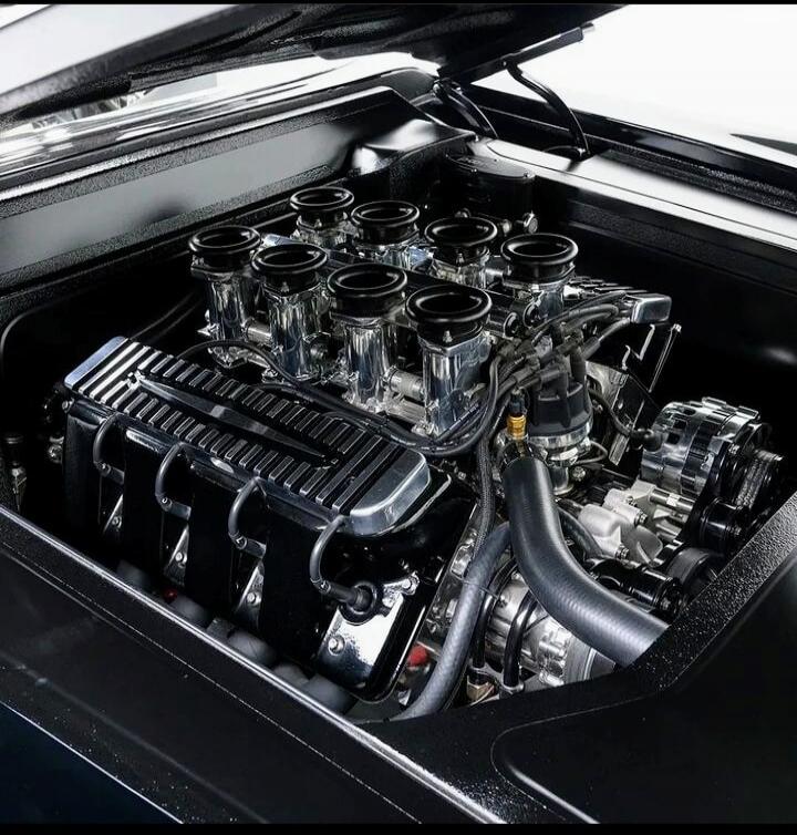 The image shows a close-up view of a powerful V8 engine, likely from a classic car. The engine features chrome accents, individual throttle bodies, and a sleek black finish, suggesting a high-performance build. The lighting highlights the intricate details and polished surfaces of the engine components.