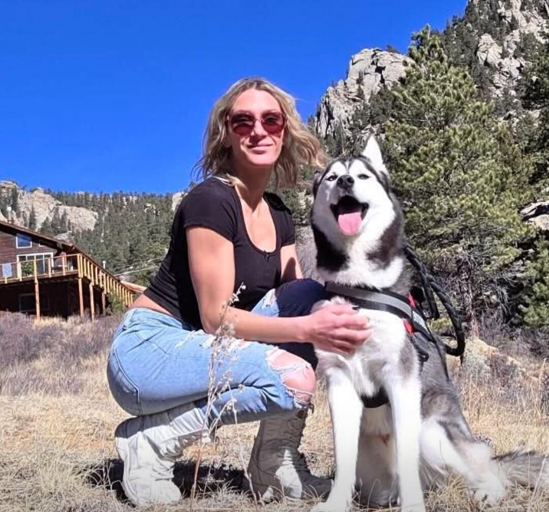 A woman with blonde hair and sunglasses is crouching next to a husky dog in a mountainous outdoor setting. Both appear happy, with the dog's tongue out and the woman smiling. There are pine trees and a house in the background under a clear blue sky.