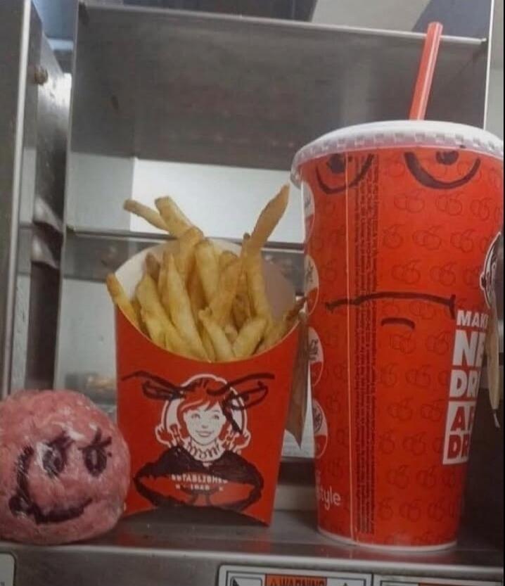 Fries in a red container with a cartoon figure printed on it, a red cup with straw, and a pink round object with a drawn face.