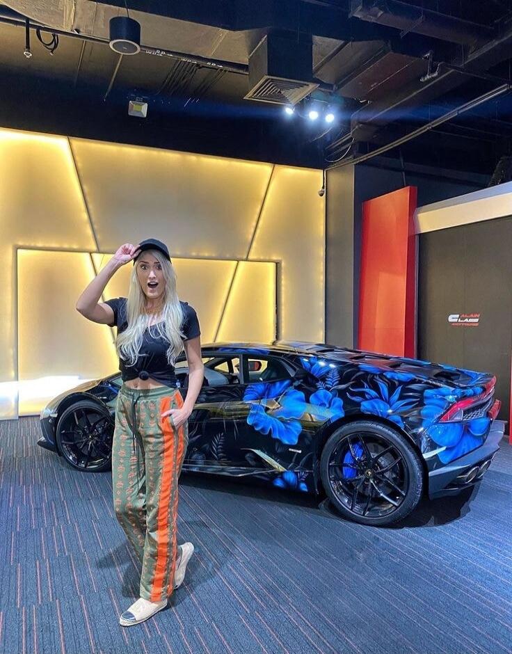 A woman with blonde hair and a black cap stands next to a black Lamborghini with a custom blue floral paint job. She has a surprised or excited expression, with her mouth open and one hand on her cap.