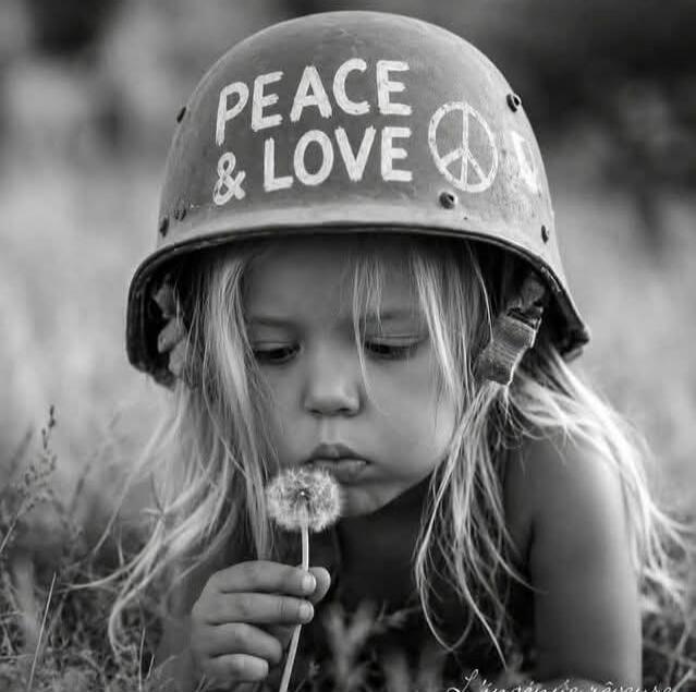 A young child with long blonde hair wears an oversized military helmet with 'PEACE & LOVE' and a peace symbol painted on it. The child is gently blowing on a dandelion in a black and white photograph.