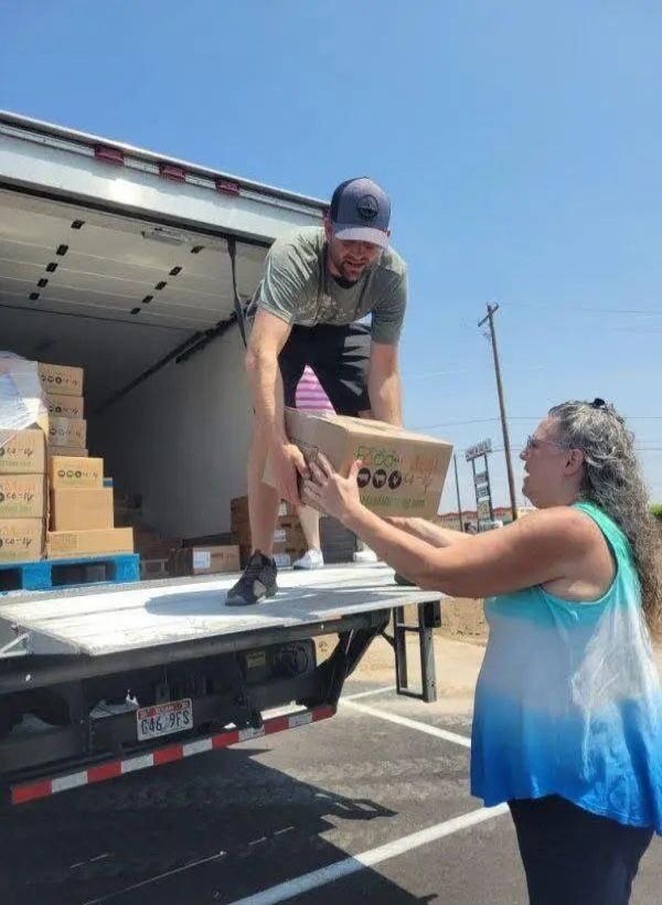 A man is unloading boxes from a truck, handing one to a woman. The boxes have 'FOOD' and '000' printed on them. A license plate '646 9FS' is visible on the truck.