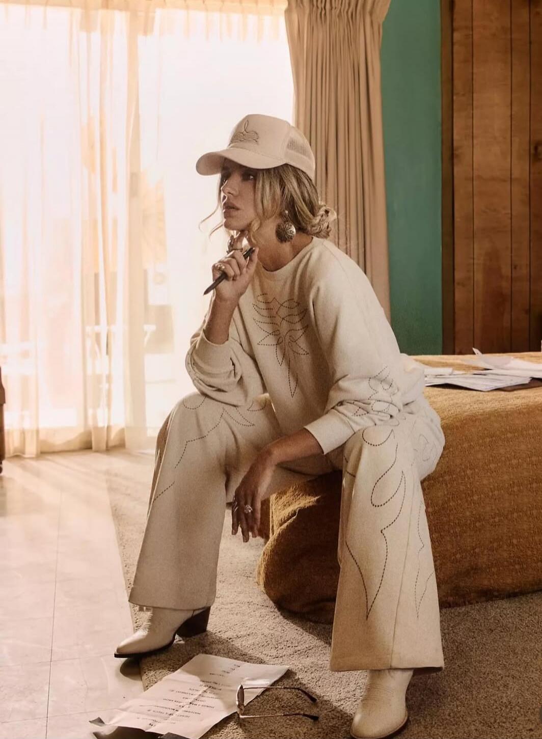 A woman in a cream-toned outfit and cap sits indoors, smoking a cigarette.
