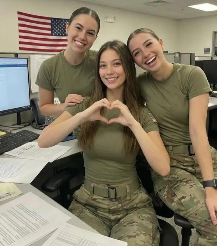 Three young women in military-style uniforms smiling in an office setting. One woman in the center is making a heart shape with her hands. An American flag is visible in the background.
