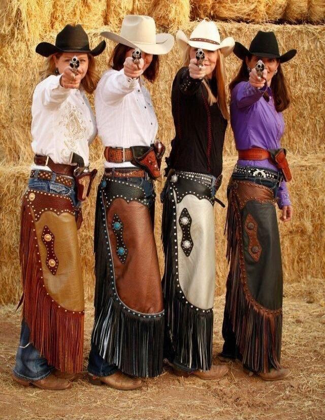 Four cowgirls pointing pistols at the camera, wearing wide-brim hats and fringed Western skirts.