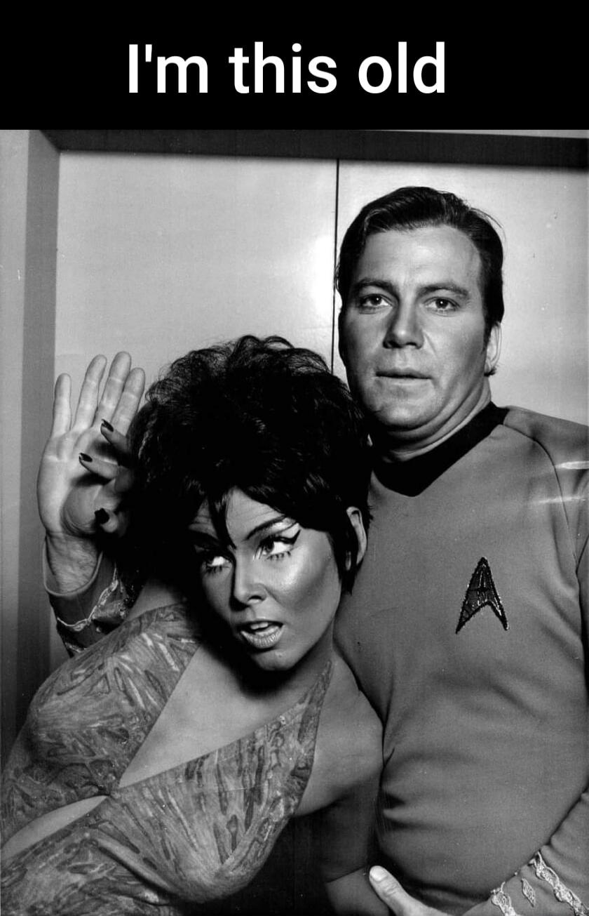 I'm this old. A man in a Starfleet uniform, resembling Captain Kirk, is standing next to a woman with elaborate makeup and a dramatic expression, who is holding up her hand.
