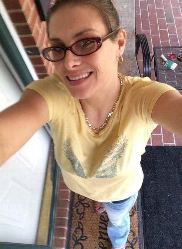 A woman smiling and taking a selfie from a high angle. She is wearing glasses, a yellow t-shirt, and a necklace. A doormat with 'Bienvenue' is visible below her.