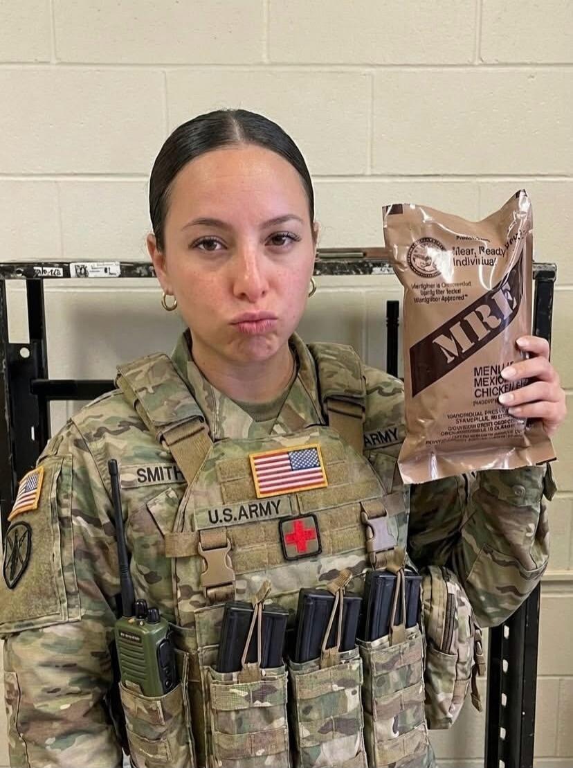 A female soldier in camouflage holding a bag labeled MRE and making a pouty face. The uniform shows U.S. Army patches and a name tag (Smith).