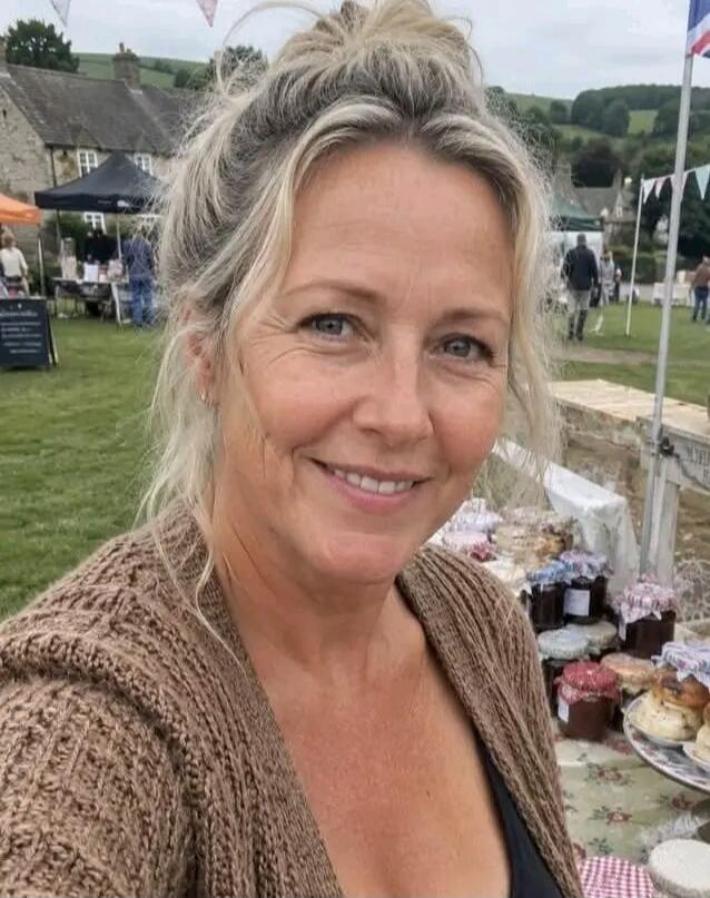 A smiling blonde woman in a brown cardigan takes a selfie at an outdoor market, with stalls selling jars and baked goods in the background.