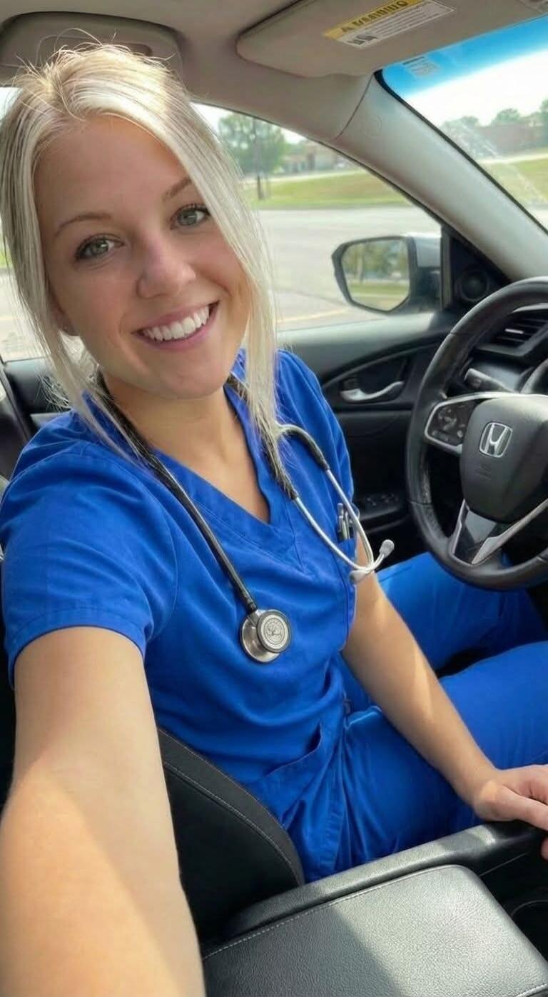 A young blonde woman in blue scrubs and a stethoscope is smiling at the camera while sitting in a car. A 'WARNING' label is visible on the sun visor.