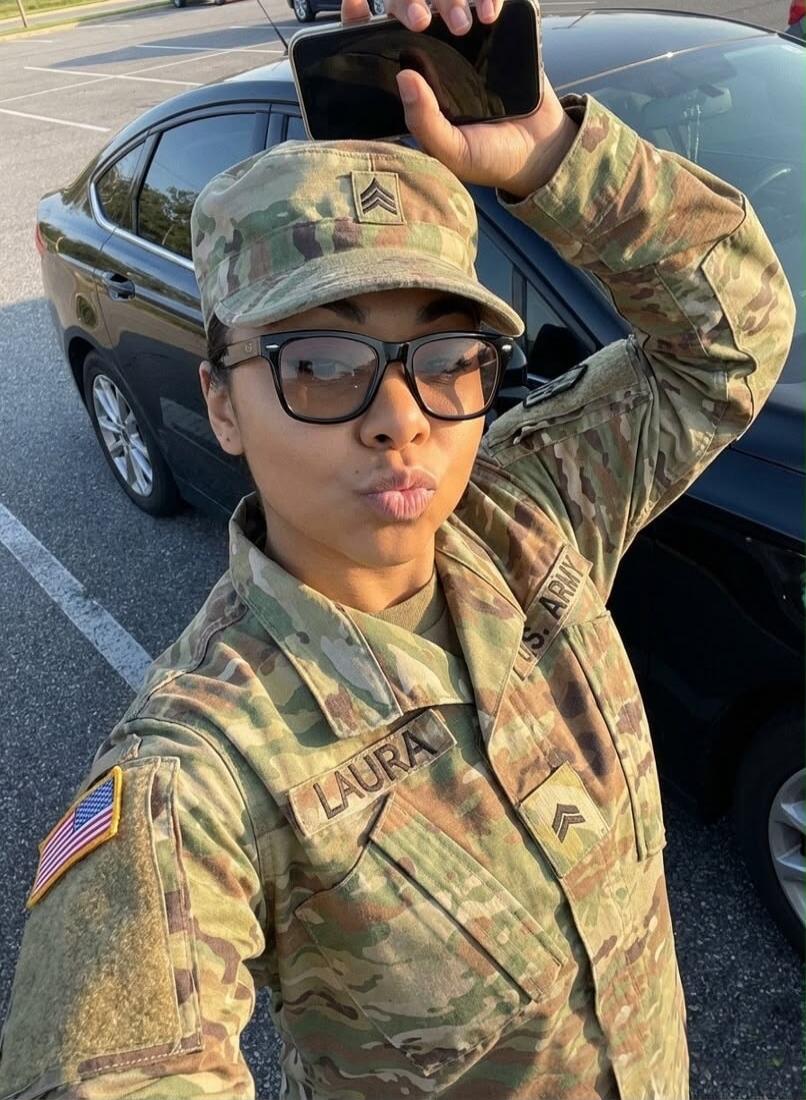 A female soldier in a U.S. Army camouflage uniform, with a name tag 'LAURA' and Staff Sergeant rank insignia, takes a selfie while making a kissing face. A US flag patch is visible on her sleeve.