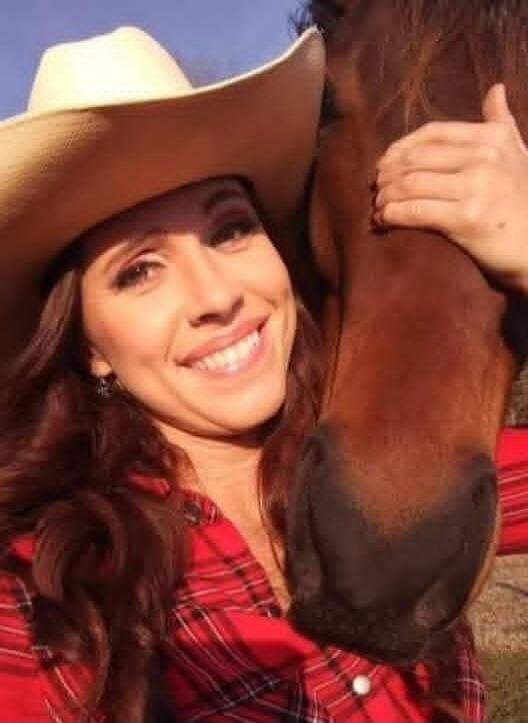 A woman in a cowboy hat and a plaid shirt is smiling next to a horse, with her arm around its neck.