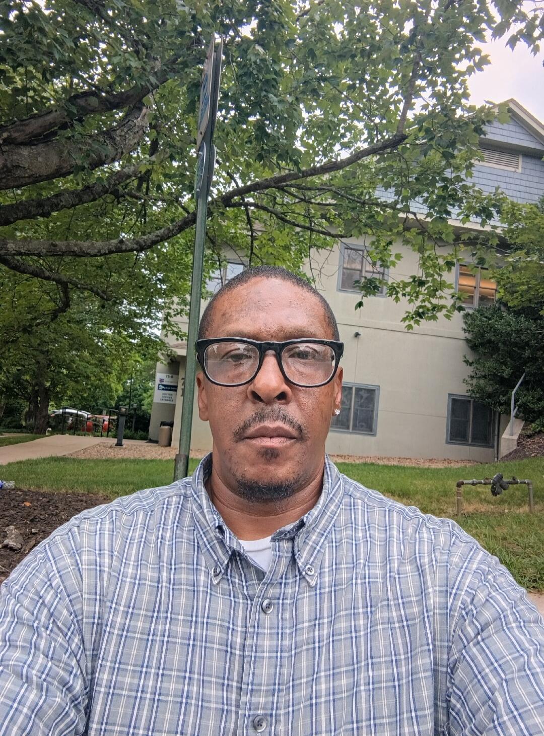 A man in a plaid shirt takes a selfie outdoors. In the background, there are trees, grass, and buildings. A sign partially visible reads '73-B' and 'ENTRANCE'.