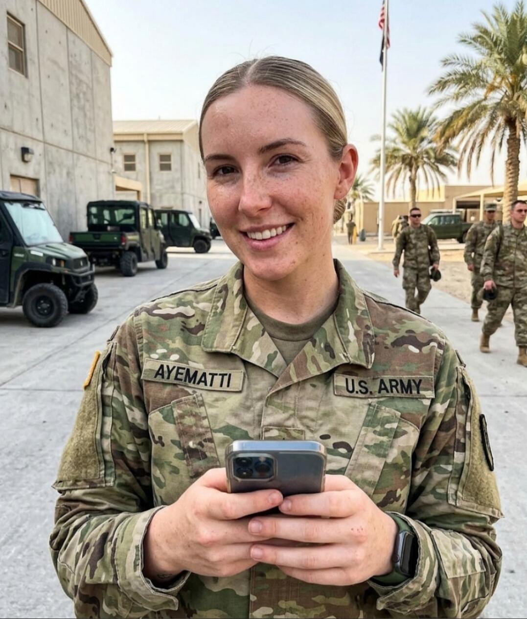 A smiling woman in a U.S. Army uniform, with a name tag 'AYEMATTI', holding a smartphone. She is outdoors on what appears to be a military base, with other soldiers, vehicles, palm trees, and a flag pole in the background.