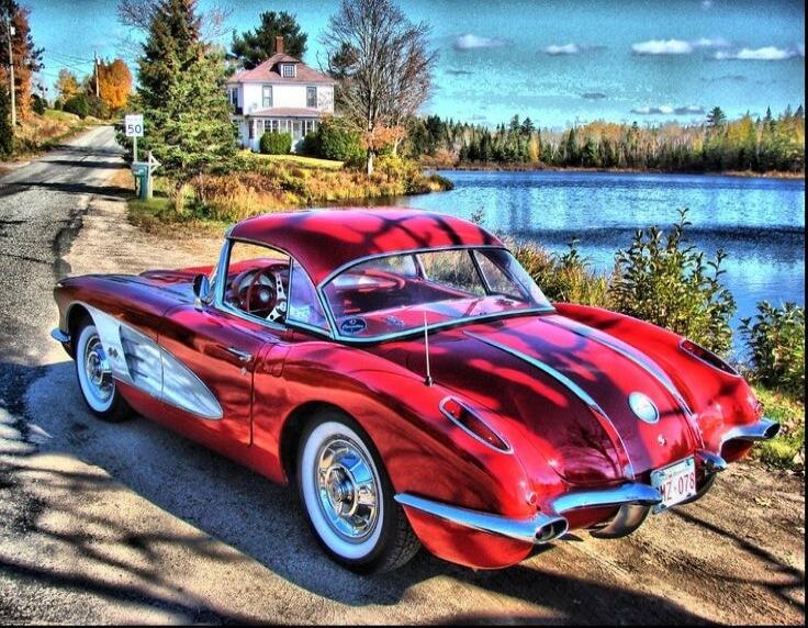 A classic red Chevrolet Corvette (C1 generation) is parked on a dirt road next to a serene lake. In the background, a white house is visible among trees under a clear blue sky. A speed limit sign '50' and a license plate 'MZ-078' are also present.
