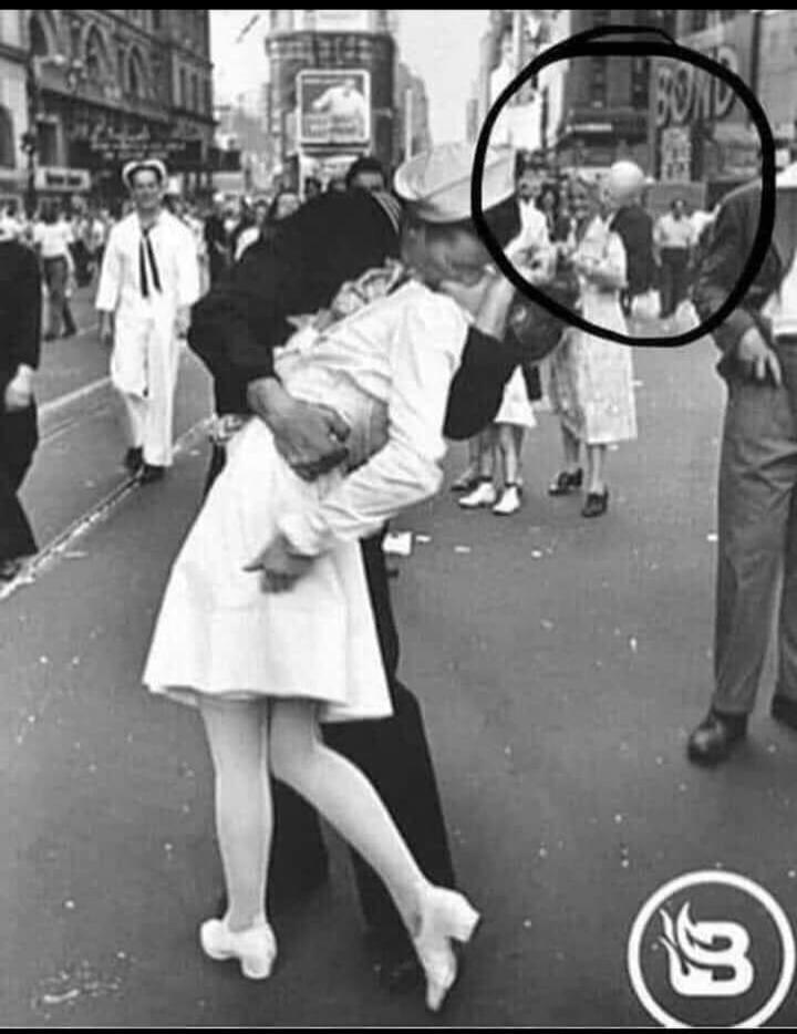 A sailor in uniform is kissing a nurse in a white dress, bending her backwards, in the middle of a street. People are visible in the background. A sign with 'BOND' is circled in the upper right.