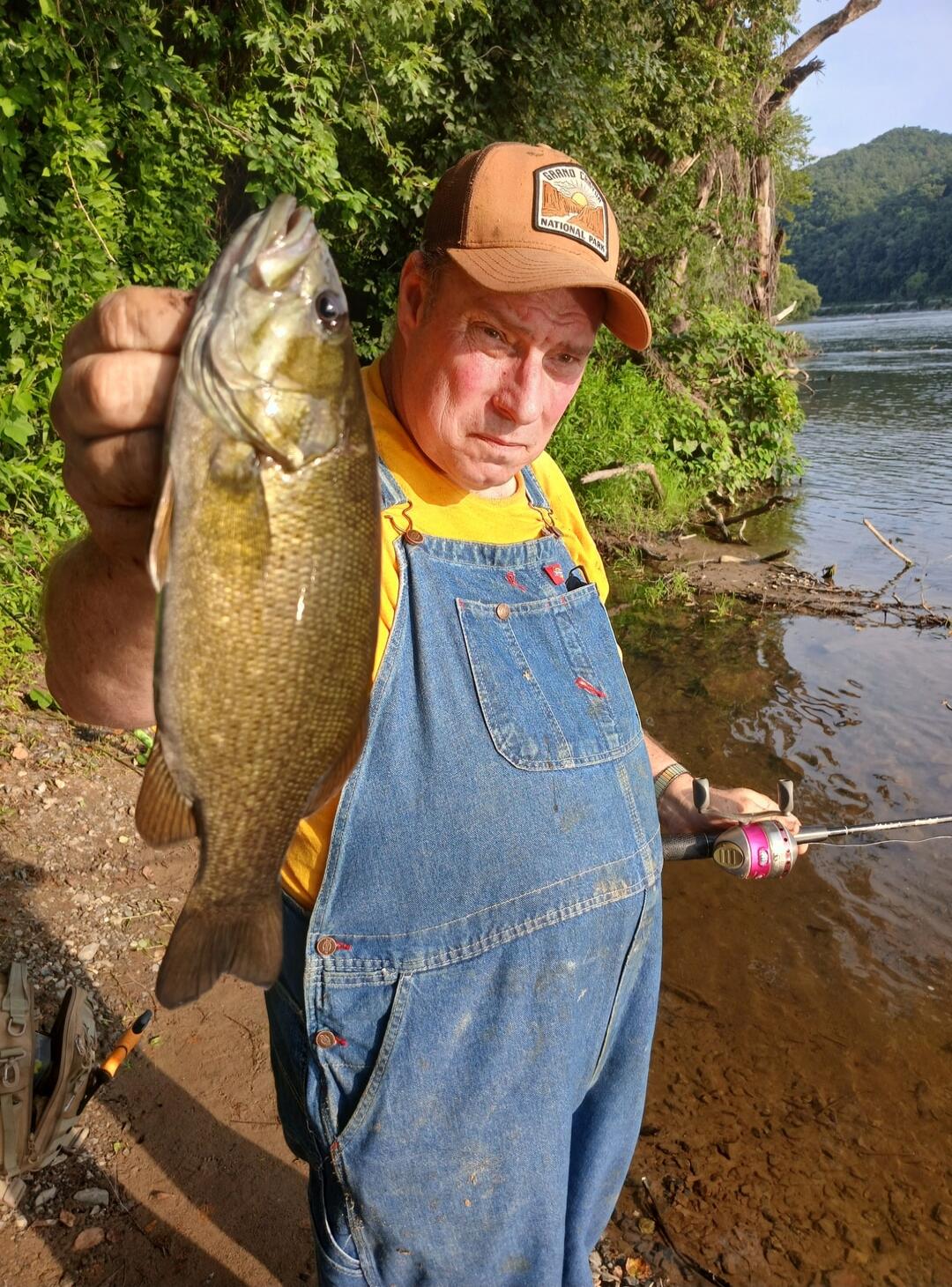 A man in a yellow shirt and blue overalls holds a freshly caught fish, possibly a bass, by a river. He is wearing a brown cap with 'GRAND CANYON NATIONAL PARK' written on it. A fishing rod is visible in his other hand. The background shows lush green trees and hills.