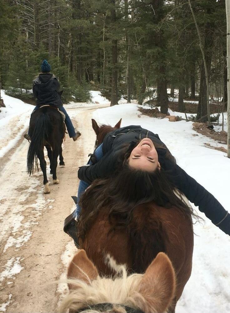 A person is riding a horse on a snowy forest path. In the foreground, the rider is laying backward over the horse's neck with arms outstretched, smiling. Another rider on a horse is seen ahead along the path.