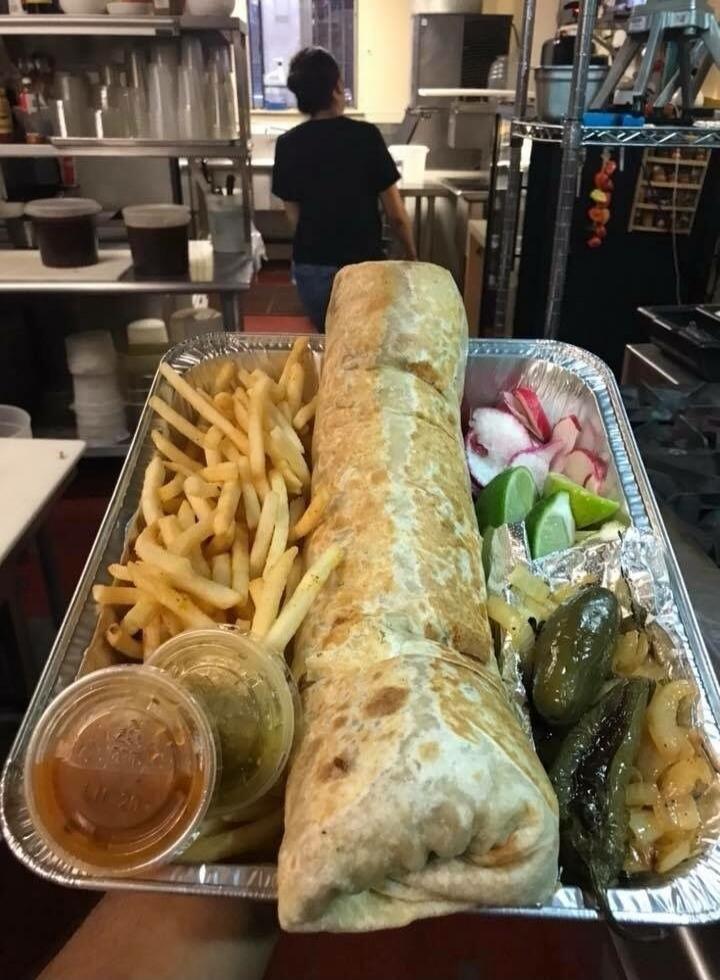 A large burrito served in a foil tray with french fries on the side. The tray also contains a small cup of sauce, lime wedges, pickles, sliced onions, and some roasted peppers or green peppers.