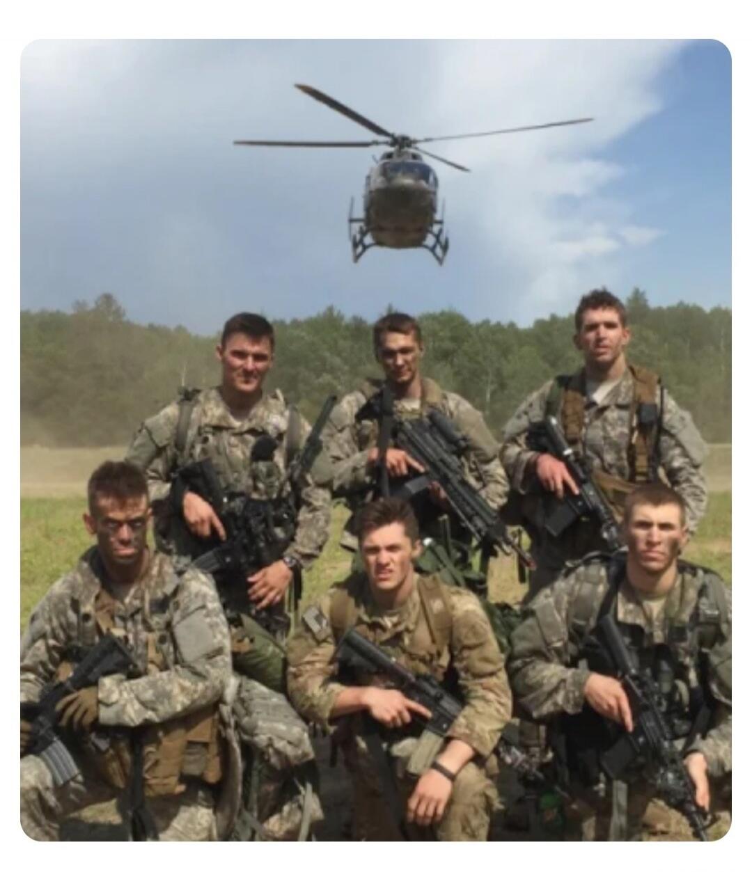 A group of six soldiers in camouflage uniforms with firearms, posing in a field with a helicopter in the background.