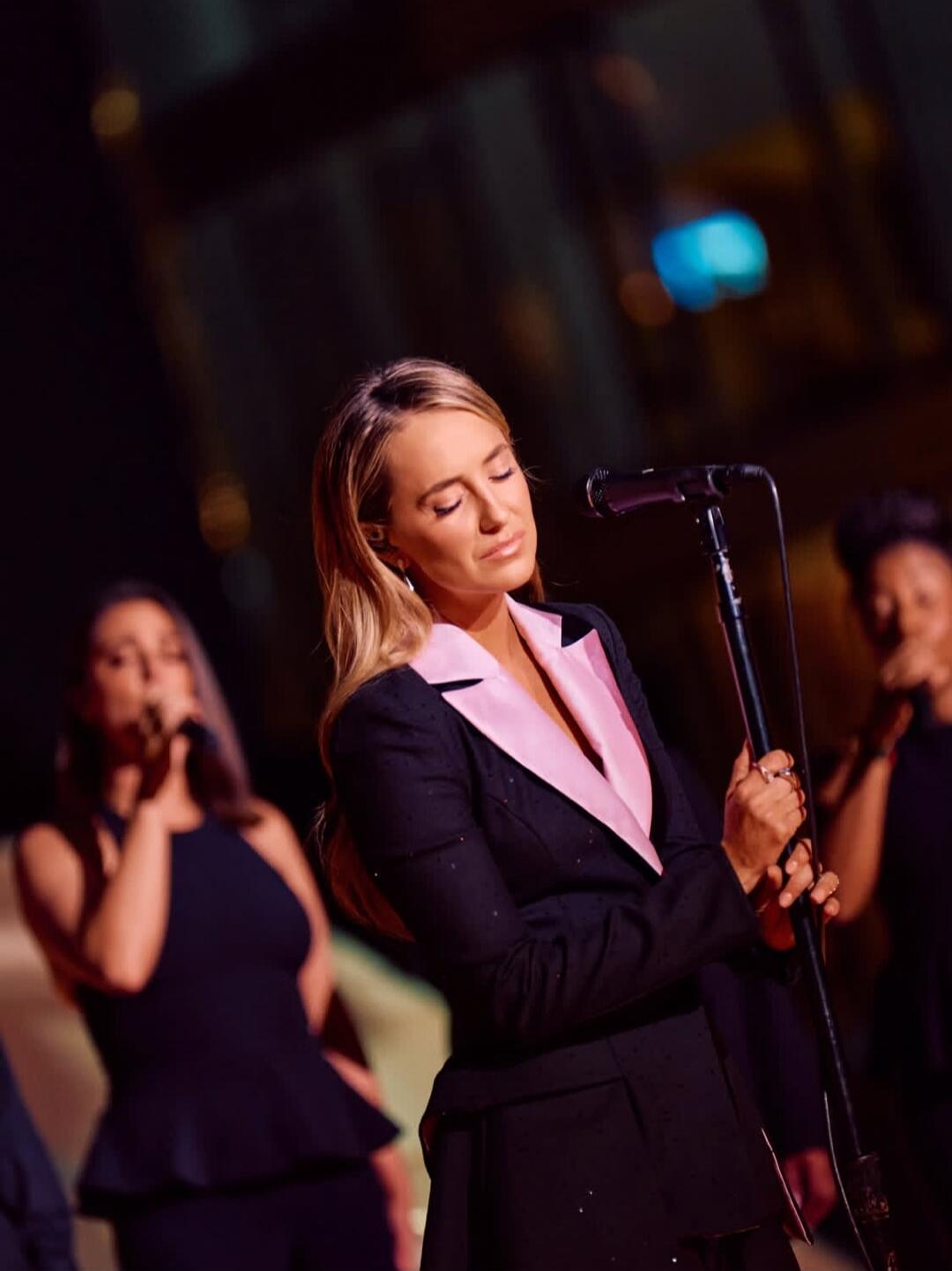 A woman with blonde hair, wearing a black suit with pink lapels, is singing into a microphone on stage with her eyes closed. Other singers are visible in the background.