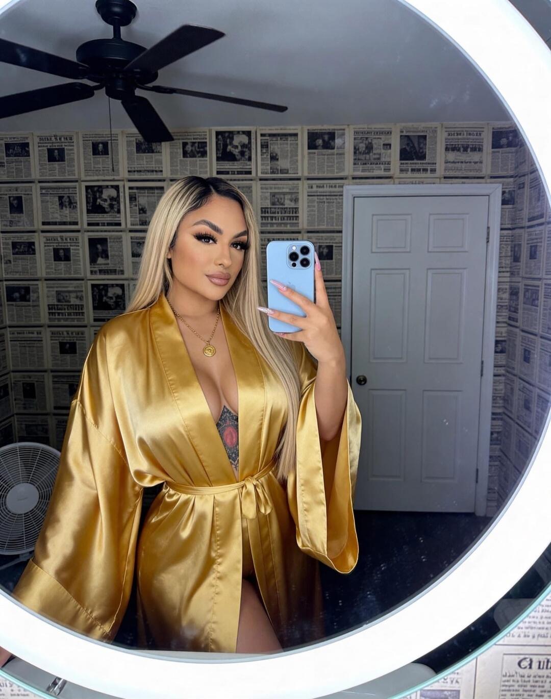 A woman in a gold satin robe taking a mirror selfie. She stands in a room with newspaper-pattern wallpaper and a circular mirror ring light; ceiling fan above. She has long blonde hair and a pendant necklace, holding a blue smartphone.