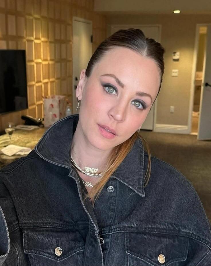 A woman wearing a dark denim jacket and a necklace with 'MILOV' text, looking directly at the camera. Her hair is pulled back, and she has light makeup.