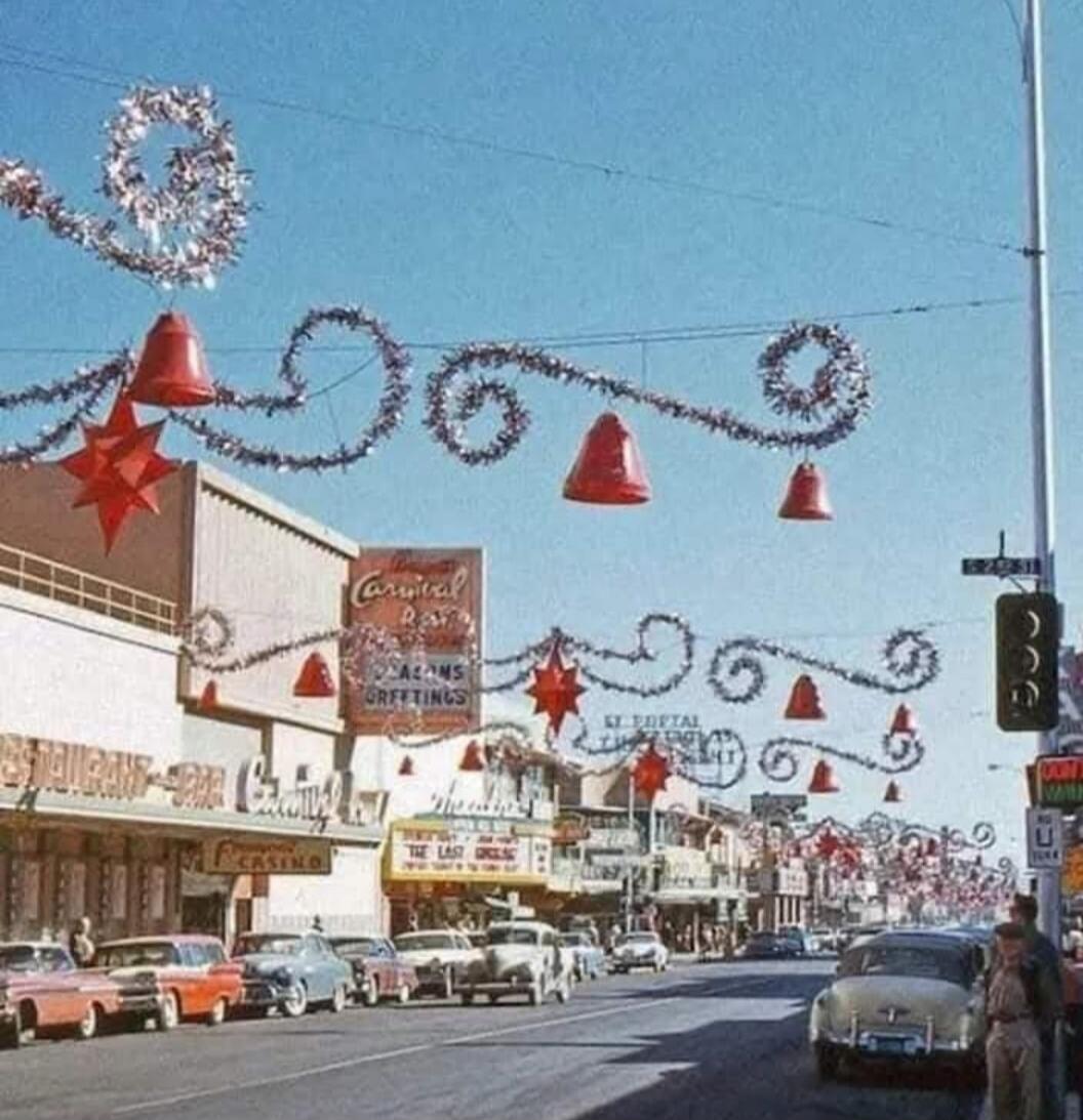 A festive street scene with decorative hanging garlands and large red star ornaments across the road. Vintage cars parked along the curb, storefronts with signs, a traffic light, and blue sky overhead.