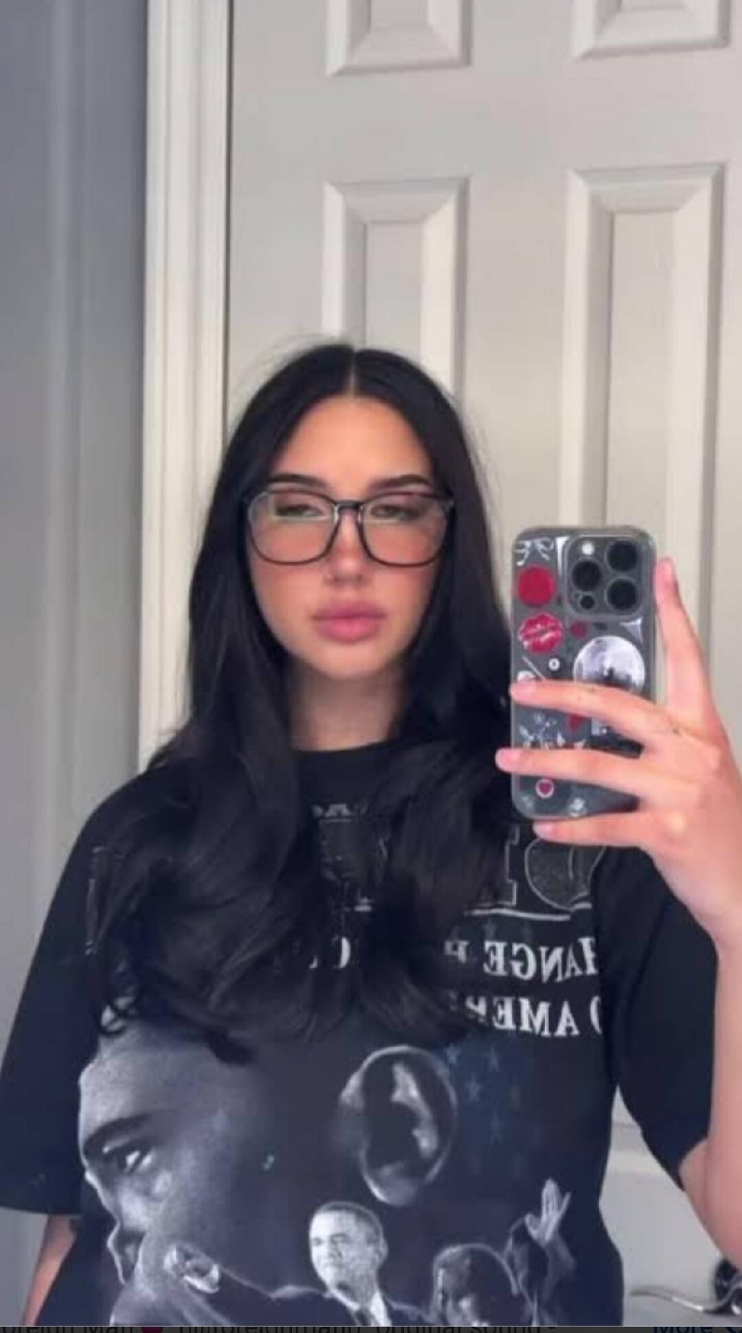 A young woman with dark hair and glasses takes a mirror selfie, holding a phone with a decorative case. She is wearing a dark t-shirt with a graphic print.