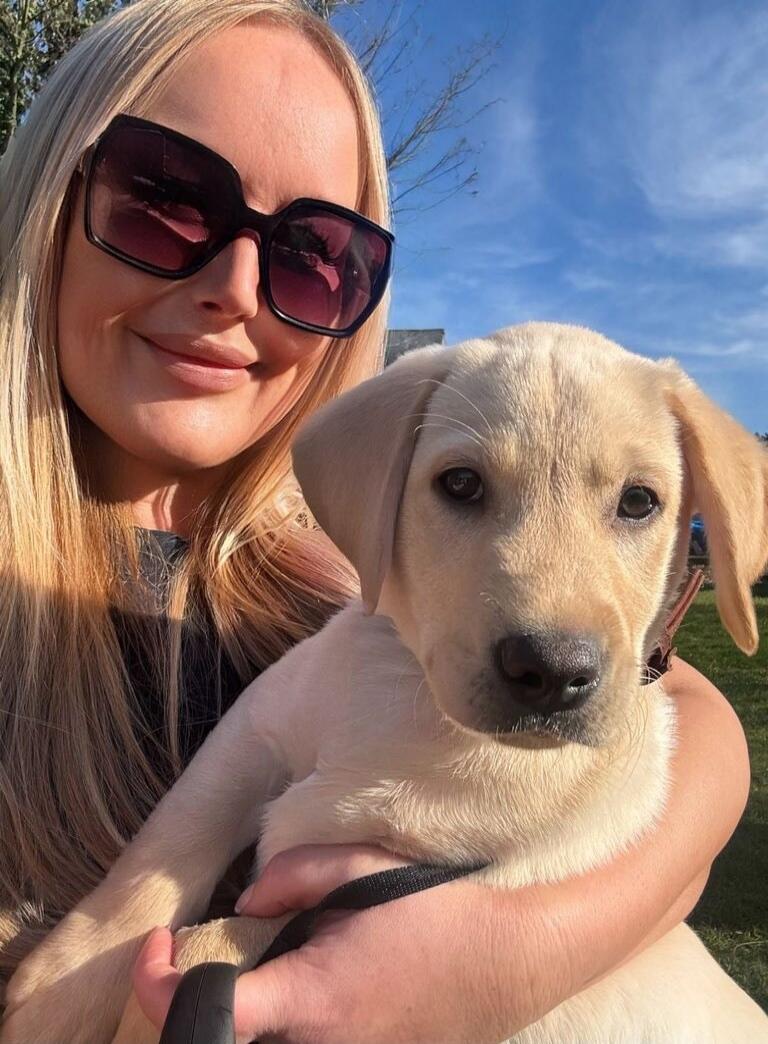 A woman with long blonde hair wearing large dark sunglasses is smiling while holding a light-colored puppy. They are outdoors on a sunny day.