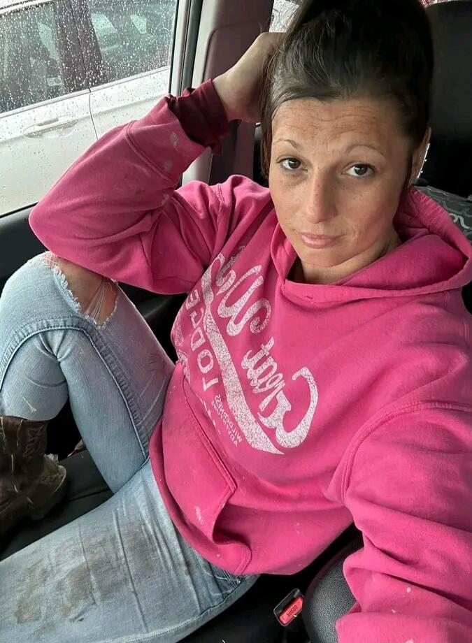 A woman in a pink hoodie and ripped jeans is sitting in a car, looking at the camera. The text on her hoodie is mirrored and unreadable.