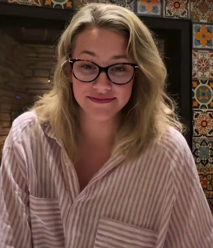 A young blonde woman with glasses is smiling slightly at the camera. She is wearing a light-colored striped shirt. The background features decorative tiles.
