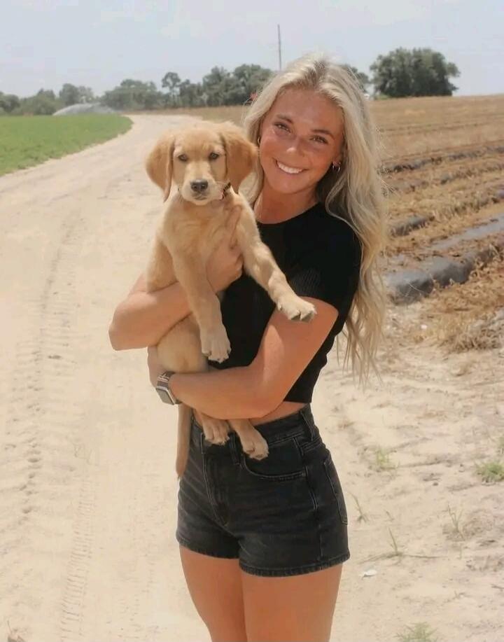 A young blonde woman is smiling and holding a golden retriever puppy on a dirt path with fields in the background. She is wearing a black crop top and denim shorts.