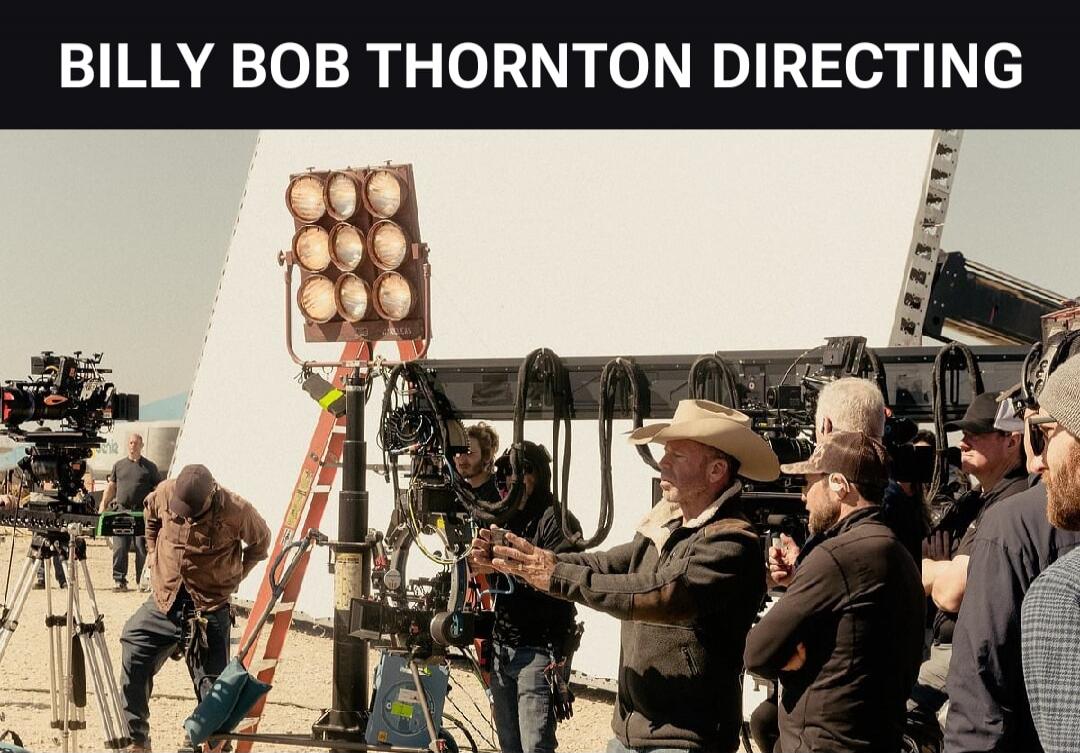 BILLY BOB THORNTON DIRECTING. A scene from a film set where Billy Bob Thornton, wearing a cowboy hat, is directing a crew. Various film equipment like cameras, lights, and reflectors are visible in an outdoor, possibly desert, location.