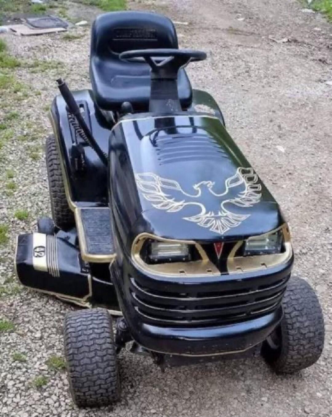 A black lawnmower customized with gold accents and a large golden phoenix decal on the hood, resembling the iconic Pontiac Trans Am design. The front grille and headlights are also styled to mimic the car's appearance. The lawnmower is parked on a gravel surface.