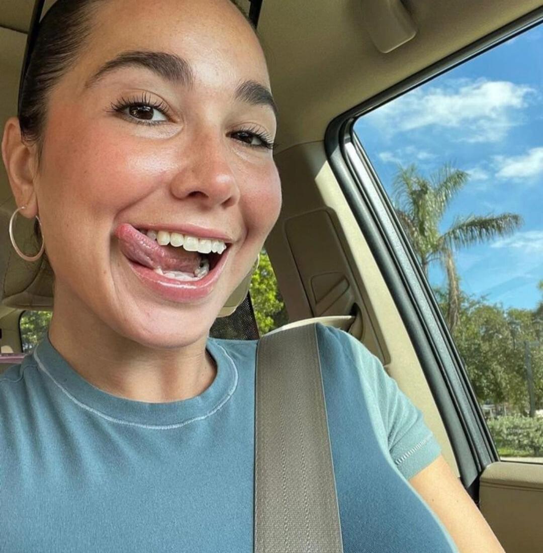 A young woman in a car, smiling broadly and playfully sticking her tongue out. She is wearing a light blue t-shirt and a seatbelt. A sunny sky with a palm tree is visible outside the window.