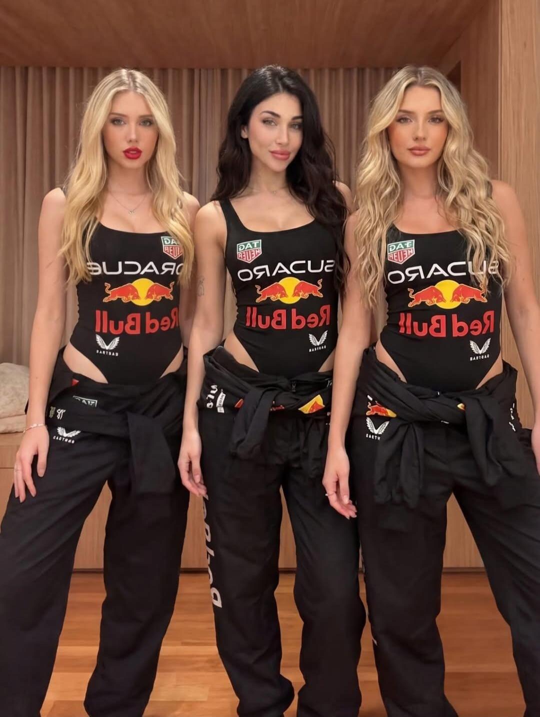 Three women wearing black tank tops with Red Bull logos, standing indoors in matching black pants with jackets tied at the waist.