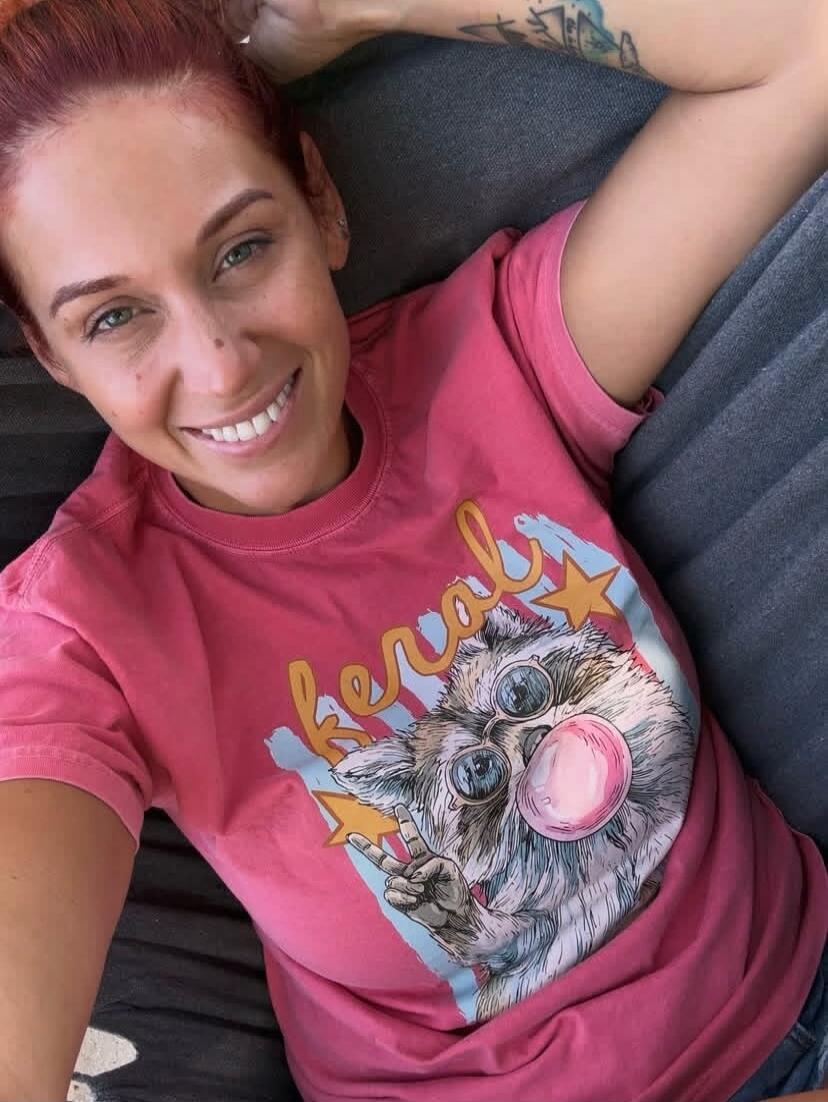 A woman with red hair and green eyes is smiling at the camera, lying down. She is wearing a pink t-shirt with a graphic of a fluffy animal wearing sunglasses, blowing a bubblegum bubble, and making a peace sign. The word 'Keral' is visible on the shirt. She has a tattoo on her arm.