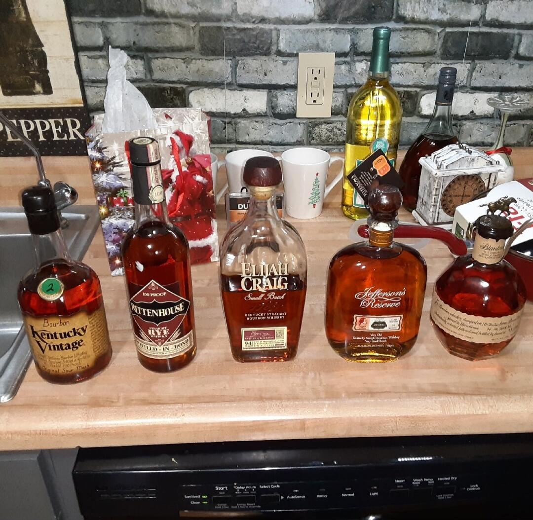 A collection of whiskey/bourbon bottles lined up on a kitchen counter. Visible labels include Elijah Craig and other bourbon brands, with a background featuring a brick-style backsplash and various kitchen items.