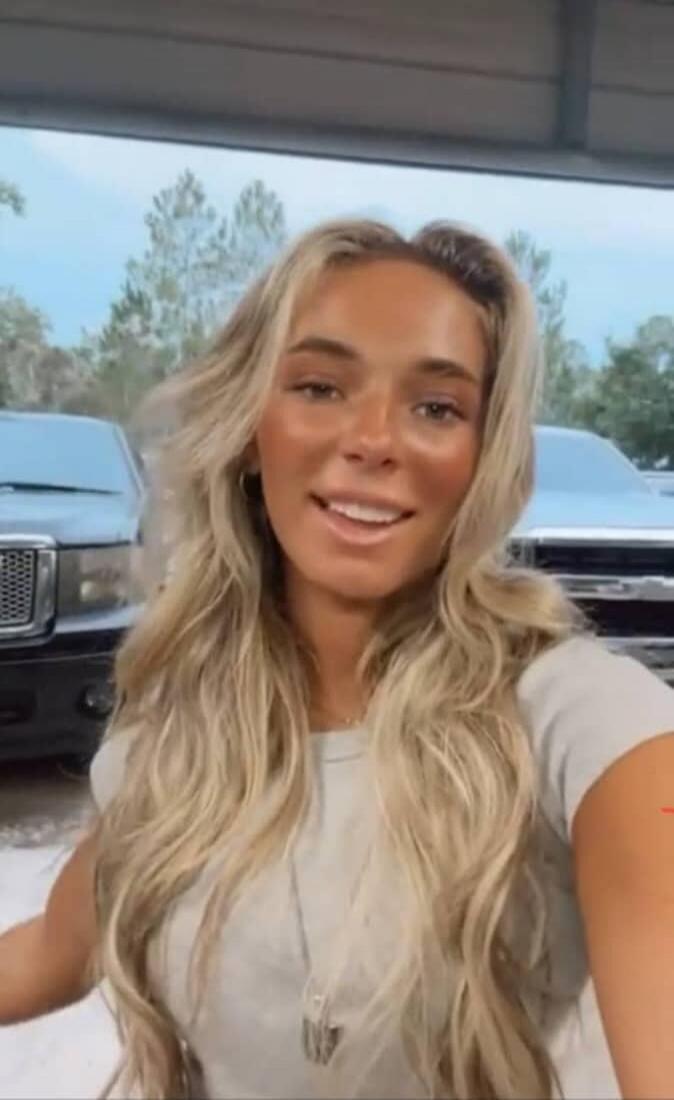A young woman with blonde hair smiles at the camera. Two pickup trucks are visible in the background.