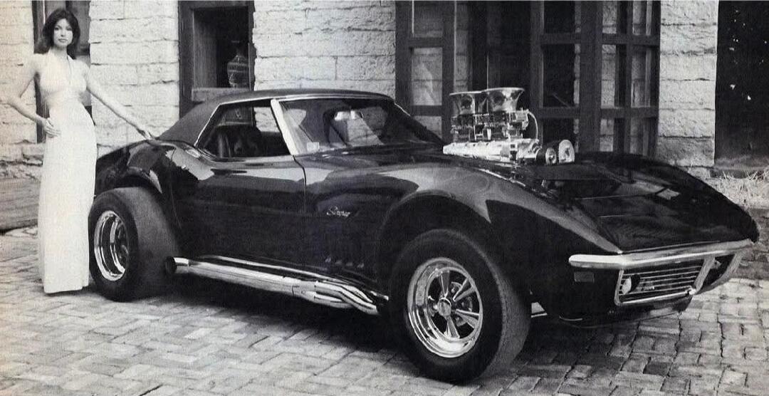 A woman in a long dress stands next to a customized classic car, possibly a Corvette, with a large engine blower visible on the hood.