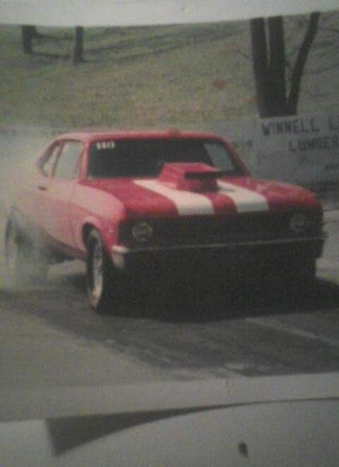 A red drag racing car with white stripes on the hood is performing a burnout, creating smoke from its rear tires. The car has the number 5813 on its windshield. In the blurry background, text possibly reads 'WINNELL L' and 'LUMBER'.
