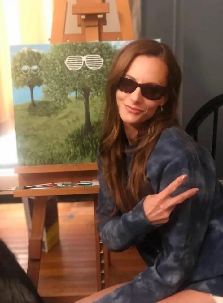 A woman in sunglasses and a tie-dye sweatshirt makes a peace sign in front of a painting of trees wearing sunglasses.