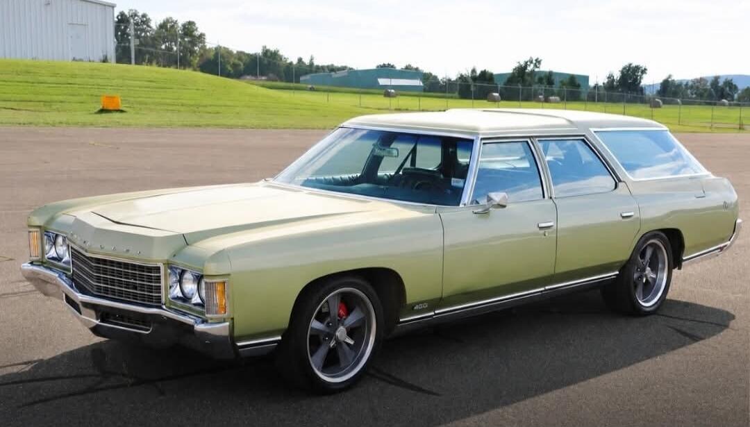 A light green vintage station wagon, possibly a Chevrolet Impala or Caprice, with a '400' badge on the side, custom wheels, and red brake calipers, parked on an asphalt surface with a grassy hill and buildings in the background.