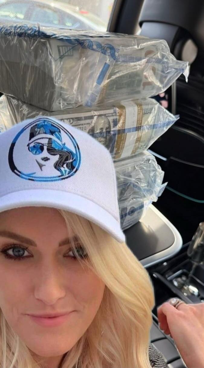 A blonde woman wearing a white cap with a blue logo takes a selfie in a car. In the background, several stacks of US hundred-dollar bills wrapped in plastic are visible.