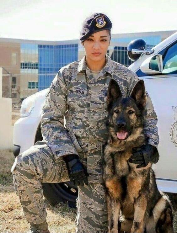 A woman in a military uniform, identified by the name tag 'HUDSON', kneels next to a German Shepherd dog. They are outdoors, with a white vehicle and a modern building in the background.
